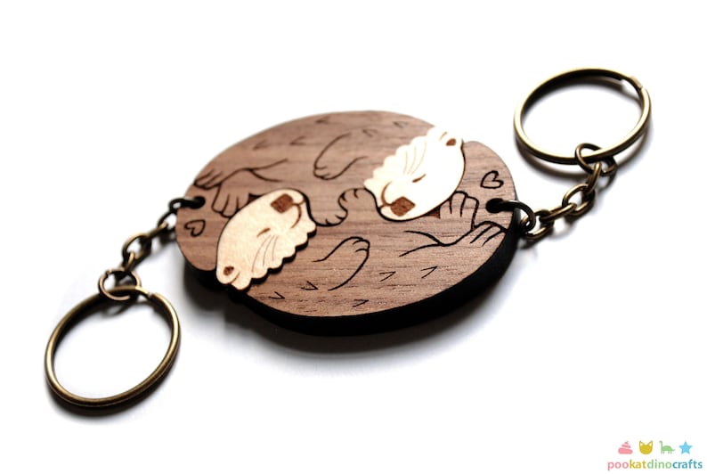 May include: Wooden keychain featuring two otters cuddling, carved in a light brown colour with a darker brown outline. The otters are holding hearts and the keychain has a bronze metal ring.