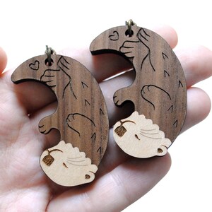 May include: A pair of wooden earrings featuring a brown and white otter design. The otters are holding hearts and are facing each other. The earrings are made of wood and have a natural finish.