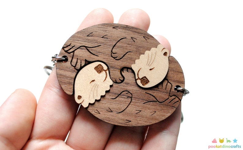 May include: A wooden keychain featuring two otters holding onto each other. The otters are carved from walnut wood and have a natural wood finish. The keychain is attached to a silver metal chain.