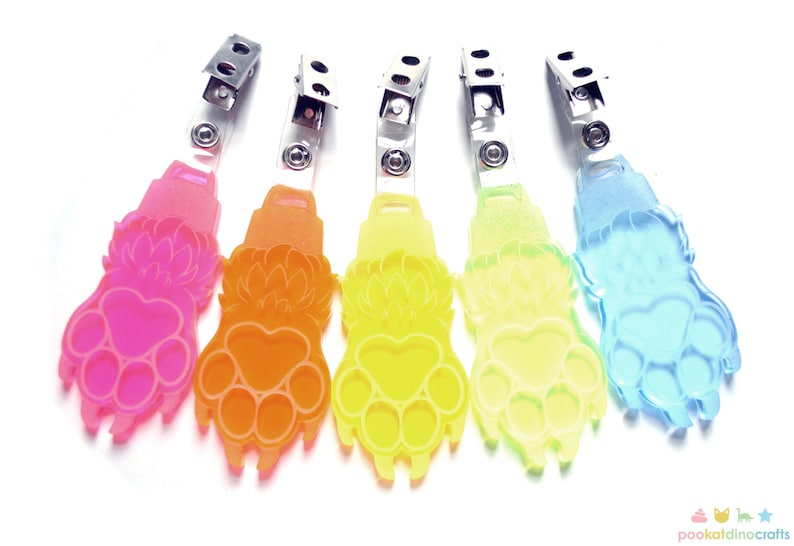 May include: Five colorful plastic paw print keychains with metal clips. The paw prints are in shades of pink, orange, yellow, light yellow, and blue.
