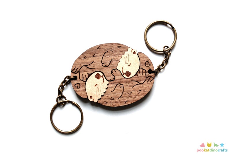 May include: Wooden keychain featuring two otters holding paws, carved in a light brown wood with a darker brown wood grain. The otters have white faces and are holding hearts. The keychain has a bronze metal ring and chain.