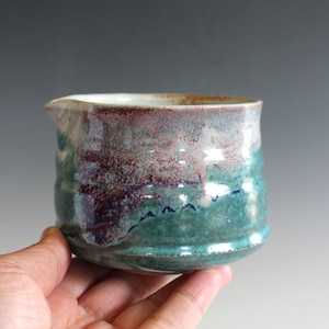 May include: A small ceramic creamer or pouring vessel with a rounded body and a small spout. The exterior features a reactive glaze with blended tones of deep teal, forest green, and dark purple, with a smooth, glossy finish and a light interior.