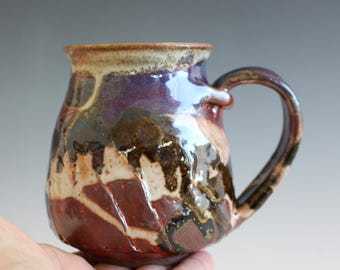 May include: A ceramic mug featuring a rounded body and a curved handle. The surface displays a multi-colored glaze with overlapping layers of deep purple, dark brown, cream, and reddish-orange tones, creating a textured, glossy appearance.