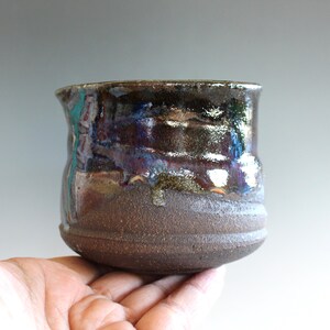May include: A small, round ceramic cup with a textured, dark brown base and a glossy, multi-colored upper glaze. The glaze features deep tones of black, metallic bronze, and streaks of iridescent blue and purple that drip down the sides.