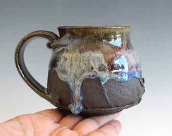 May include: A handmade ceramic mug with a dark brown base and a handle. The top of the mug has a glossy glaze with shades of brown, blue, and white. The mug is designed for hot beverages.