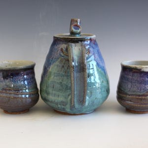 May include: A handmade ceramic tea set with a teapot and two matching cups. The teapot is a rounded shape with a handle and lid. The set features a blend of blue, green, and brown colors, creating a unique, artistic look. The cups are approximately 3 inches tall.