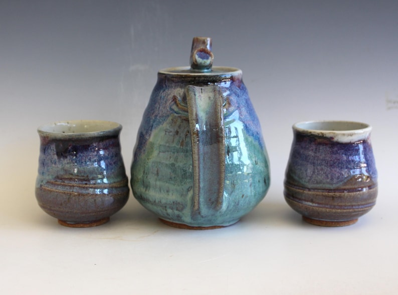 May include: A handmade ceramic tea set with a teapot and two matching cups. The teapot is a rounded shape with a handle and lid. The set features a blend of blue, green, and brown colors, creating a unique, artistic look. The cups are approximately 3 inches tall.