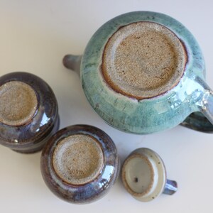May include: A ceramic tea set with a teal teapot, two small cups, and a lid. The teapot and cups have a glossy finish with shades of blue, purple, and brown. The bottoms of the pieces are unglazed.