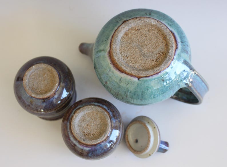 May include: A ceramic tea set with a teal teapot, two small cups, and a lid. The teapot and cups have a glossy finish with shades of blue, purple, and brown. The bottoms of the pieces are unglazed.