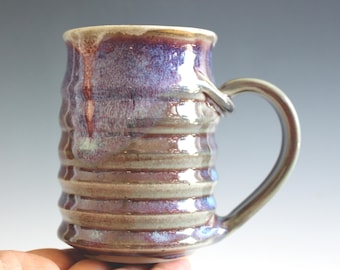 May include: A ceramic mug with a cylindrical body featuring horizontal ridges. The glaze is a blend of deep purple, blue, and earthy brown tones with a glossy finish. It has a curved handle attached to the side and a wide, circular rim at the top.