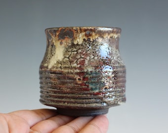 May include: A small, cylindrical ceramic cup featuring a textured, multi-colored glaze with shades of brown, cream, and hints of red. The exterior surface displays a crackled, mottled finish with horizontal ridges around the lower section.