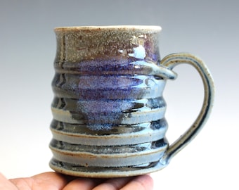 May include: Handmade ceramic mug with a unique, textured design. The mug features a blue and brown glaze with a ribbed pattern around the body. The handle is a curved loop. The mug is held in a hand.