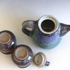 May include: A ceramic tea set with a teapot, two cups, and a lid. The teapot and cups are glazed in shades of blue, green, and brown. The teapot has a handle and spout. The lid is off to the side.