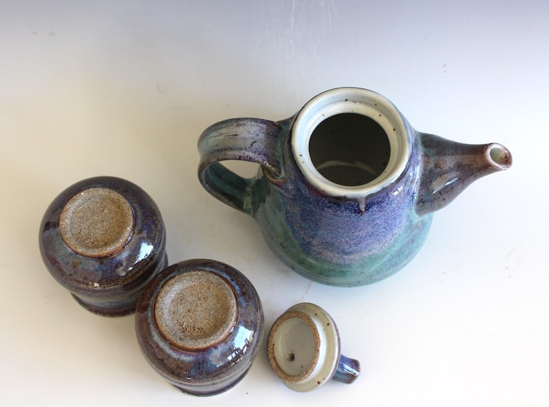 May include: A ceramic tea set with a teapot, two cups, and a lid. The teapot and cups are glazed in shades of blue, green, and brown. The teapot has a handle and spout. The lid is off to the side.