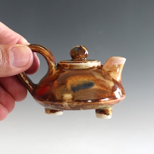 May include: A small, brown ceramic teapot with a lid and three legs. The teapot has a mottled glaze with shades of brown, blue, and red.
