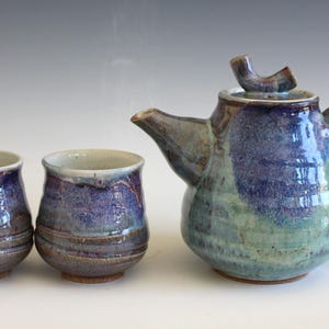 May include: A handmade ceramic tea set with a teapot and two matching cups. The teapot and cups are glazed in shades of blue, purple, and green. The teapot has a curved spout and handle. The cups are small and rounded.