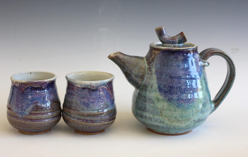 May include: A handmade ceramic tea set with a teapot and two matching cups. The teapot and cups are glazed in shades of blue, purple, and green. The teapot has a curved spout and handle. The cups are small and rounded.