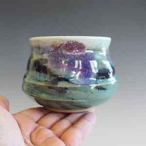 May include: A handmade ceramic cup with a rounded shape. The cup features a blend of colors, including teal, purple, and dark blue, with a textured surface. The rim is a light color, and the base is a natural clay color.