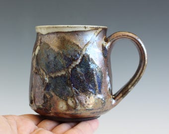 May include: A ceramic mug with a rounded body and a curved handle. The exterior features a mottled glaze with deep blue, brown, and metallic bronze tones, accented by light-colored crackle patterns and a smooth, off-white rim at the top edge.