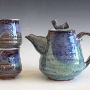May include: A handmade ceramic tea set with a teapot and two cups. The teapot and cups are glazed in shades of blue, purple, and green. The teapot has a curved handle and a spout. The cups are stacked. The set is on a white surface.