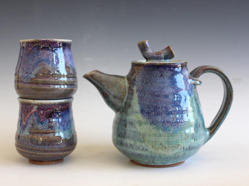 May include: A handmade ceramic tea set with a teapot and two cups. The teapot and cups are glazed in shades of blue, purple, and green. The teapot has a curved handle and a spout. The cups are stacked. The set is on a white surface.