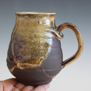 May include: Handmade ceramic mug with a textured, earthy design. The mug features a dark brown base, transitioning to a lighter brown and tan upper portion. The handle is a matching brown. The mug is held in a hand.