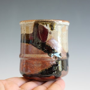 May include: A cylindrical ceramic cup featuring a variegated glaze with earthy tones of deep brown, rust red, and cream. The surface displays a glossy, dripped texture with a prominent horizontal ridge encircling the lower section of the vessel.