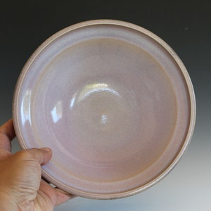 May include: A large, round, ceramic bowl with a light pink glaze. The bowl has a slightly raised rim and a smooth, glossy finish.