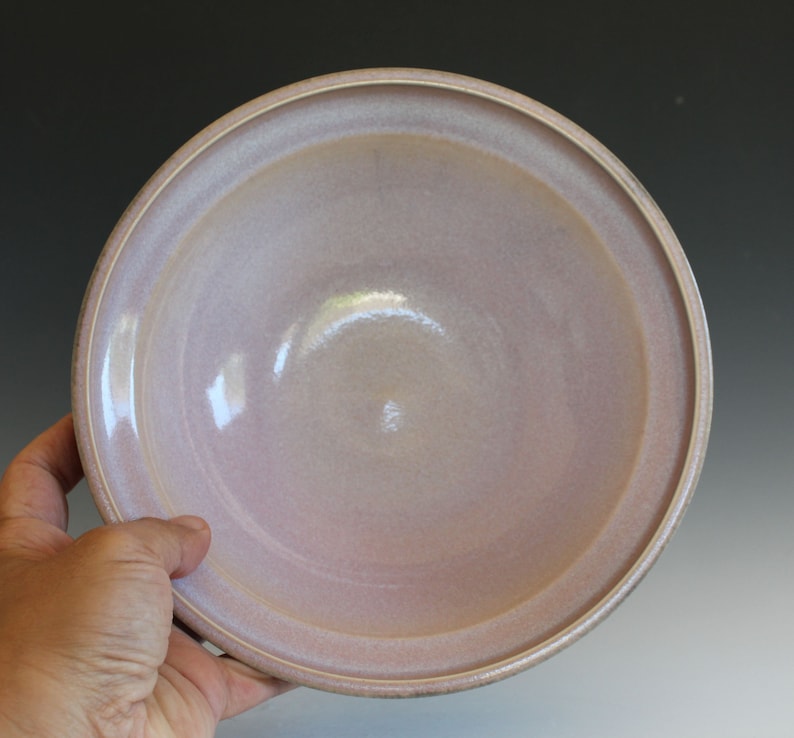 May include: A large, round, ceramic bowl with a light pink glaze. The bowl has a slightly raised rim and a smooth, glossy finish.
