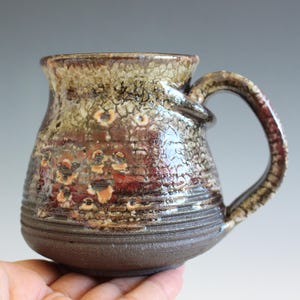May include: A ceramic mug with a wide, rounded base and a curved handle. The upper portion features a textured, crackled glaze in shades of brown, tan, and deep red, while the lower section has a dark, matte finish with horizontal ridges.