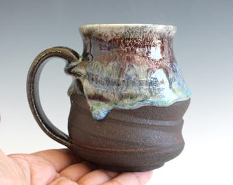 May include: A handmade ceramic mug with a dark brown base and a textured, swirling design. The upper portion features a blend of colors, including brown, blue, and white, with a glossy finish. The mug has a curved handle.