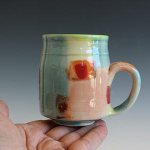 May include: A hand-painted ceramic mug with a rounded body and a curved handle. The mug features a colorful design with patches of red, orange, and blue hues. The mug is held in a person's hand.