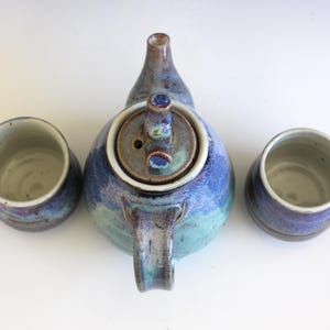 May include: A handmade ceramic tea set with a blue, teal, and purple glaze. The set includes a teapot with a lid and two matching cups. The teapot has a curved handle and spout.