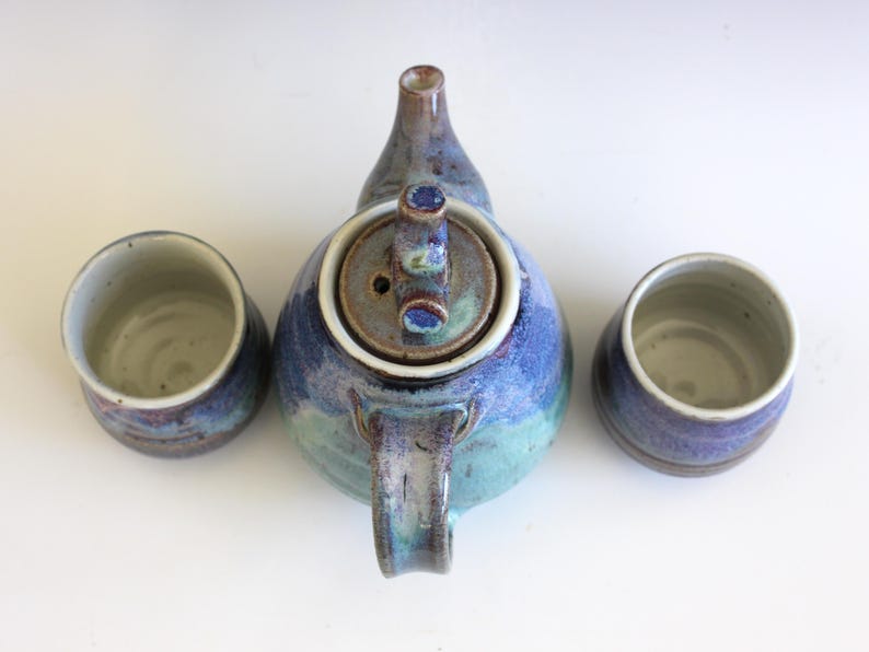 May include: A handmade ceramic tea set with a blue, teal, and purple glaze. The set includes a teapot with a lid and two matching cups. The teapot has a curved handle and spout.