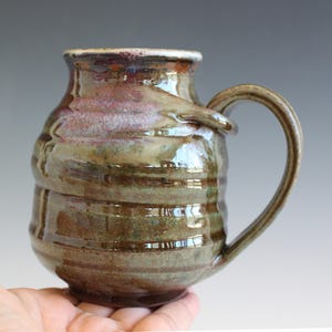 May include: A ceramic mug with a rounded body and a single curved handle. The surface features a glossy glaze in shades of olive green, brown, and hints of purple, with horizontal ridges circling the exterior and a smooth, flared rim at the top.