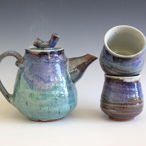May include: A handmade ceramic teapot and two matching cups. The teapot is a rounded shape with a curved handle and spout. The colors are a blend of teal, purple, and brown. The cups are stacked and have a similar color scheme.