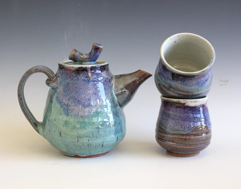 May include: A handmade ceramic teapot and two matching cups. The teapot is a rounded shape with a curved handle and spout. The colors are a blend of teal, purple, and brown. The cups are stacked and have a similar color scheme.
