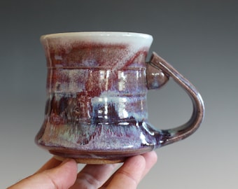 May include: A handmade ceramic mug with a unique glaze featuring shades of purple, blue, and brown. The mug has a wide base and a rounded handle.