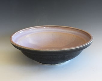 May include: A large, round ceramic bowl with a dark gray exterior and a light purple interior. The bowl has a slightly flared rim and a smooth, glazed finish.