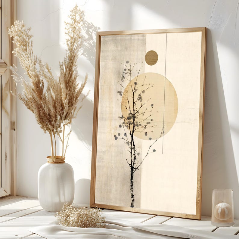 May include: A framed abstract art print featuring a tree silhouette with a large yellow circle and a smaller gold circle. The artwork has a neutral color palette with beige and cream tones. A white vase with dried pampas grass and a candle in a glass holder are in the foreground.