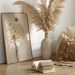 May include: A framed abstract art print with a beige and brown color scheme, featuring a tree silhouette and a sun-like shape. A large vase with pampas grass, a throw blanket, and an open book are also in the room. The room has a neutral color palette.