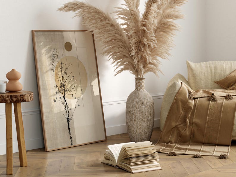 May include: A framed abstract art print with a beige and brown color scheme, featuring a tree silhouette and a sun-like shape. A large vase with pampas grass, a throw blanket, and an open book are also in the room. The room has a neutral color palette.