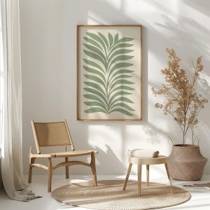 May include: A framed print with a green leaf design on a cream background. The artwork hangs above a wooden chair, a small stool, and a round jute rug. The scene evokes a sense of calm, with natural materials and a minimalist style.