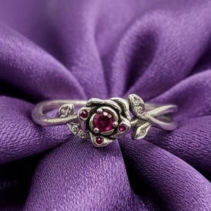 Pode incluir: Um anel de prata com um design de rosa, com uma pedra preciosa vermelha central e pequenos detalhes vermelhos. O anel tem detalhes delicados de folhas e videira, sobre um fundo de tecido roxo.