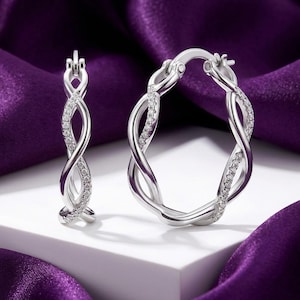 May include: A pair of silver hoop earrings featuring a twisted design with one strand encrusted with small clear stones. The earrings have a hinged closure mechanism and a polished metallic finish, showcasing a detailed intertwined structure.