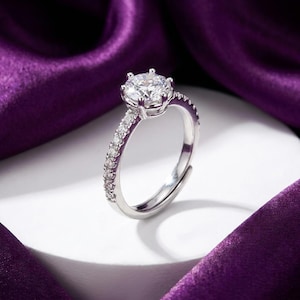 Peut inclure: Une bague argentée dotée d'une grande pierre centrale ronde et transparente à facettes, maintenue par un sertissage à six griffes. L'anneau est orné d'une rangée de petites pierres rondes transparentes sur la moitié supérieure, avec un design ouvert.
