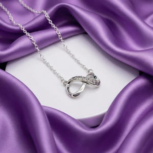 May include: A silver-toned necklace with an infinity symbol pendant, embellished with small, clear gemstones. The chain and pendant are displayed on a white surface against a backdrop of draped, vibrant purple fabric.