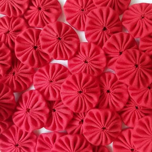 May include: A collection of red fabric rosettes, also known as  ruffle flowers, arranged in a close-up view. The rosettes are made of a soft, textured fabric and have a small hole in the center.