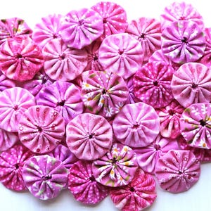May include: A collection of pink and purple fabric rosettes with various floral patterns. The rosettes are arranged in a heart shape on a white background.