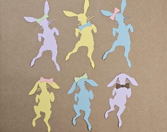 以下が含まれることがあります： 黄色、青、紫など、さまざまな色のウサギの紙切りが6つあります。各ウサギはリボンまたは蝶ネクタイをしており、すべて遊び心のあるポーズをとっています。